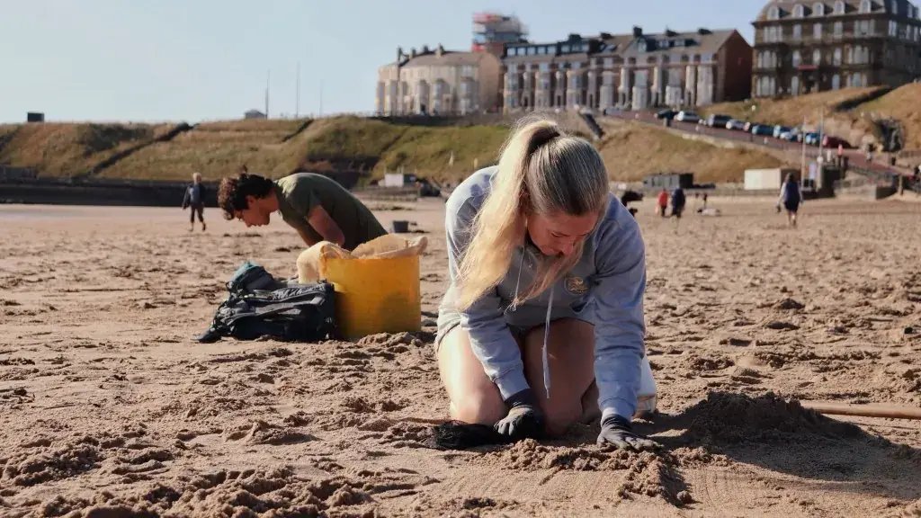 Volunteer in gloves searching sand for nurdles beside a yellow bucket