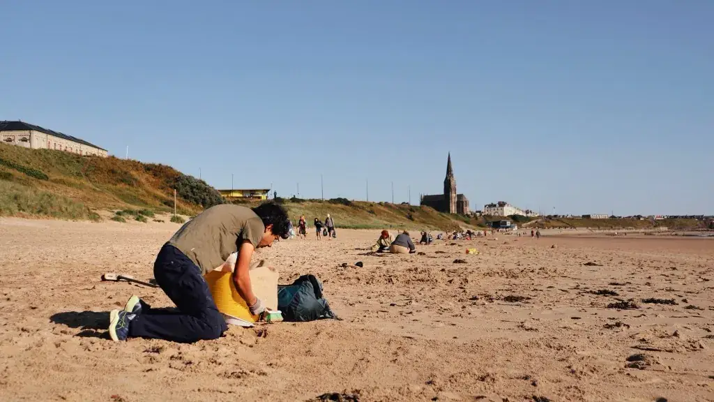 Volunteer kneeling on the beach collecting nurdles with a yellow container, St George’s Church behind