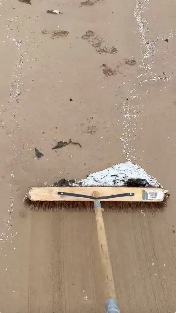 Wooden broom scraping white nurdles across wet beach sand