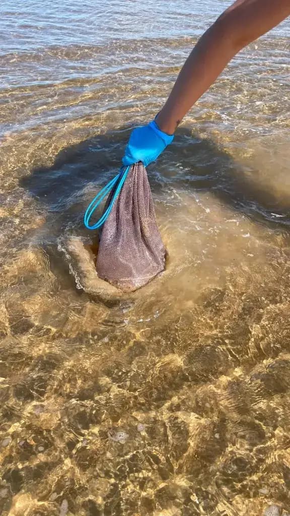 Purple mesh bag being washed in shallow seawater while wearing a blue glove